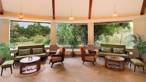 A spacious room with a high ceiling, wooden beams, and large windows offering a view of a lush garden. - The Safari Lodge Pench (Kurai)