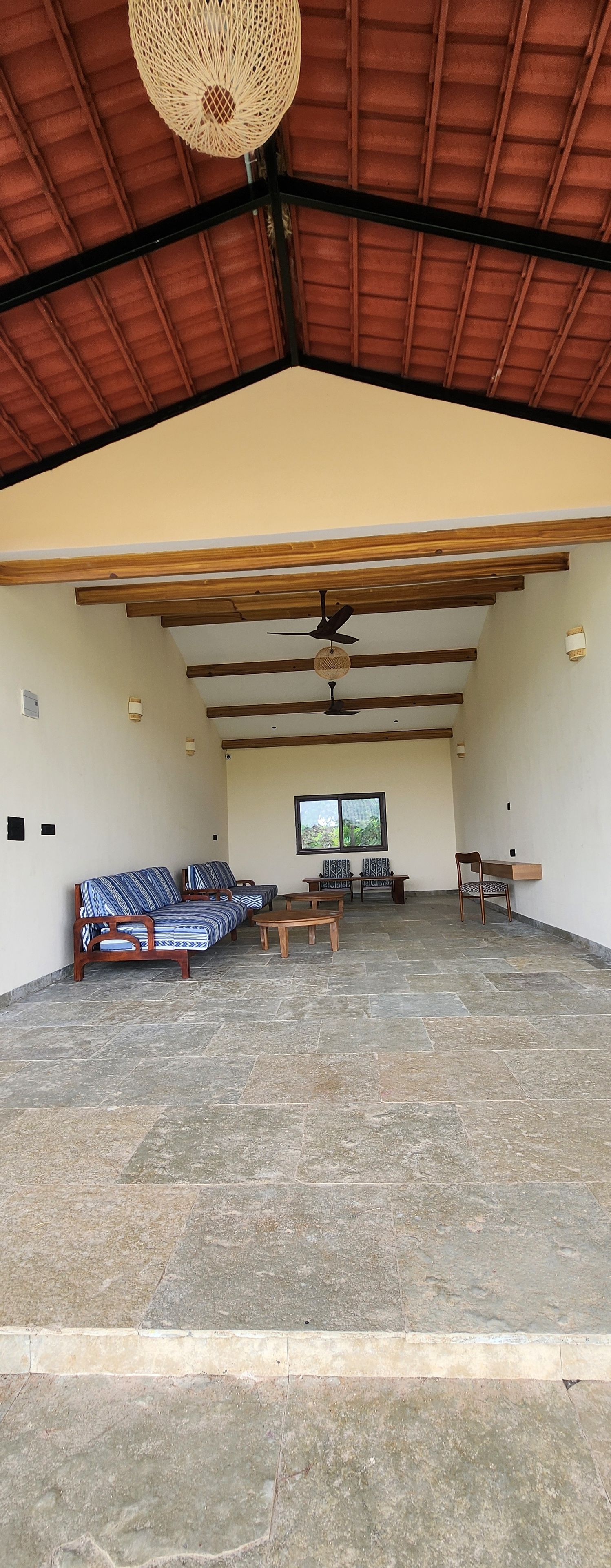 A spacious indoor area with a high ceiling, wooden beams, a ceiling fan, and a large window providing natural light.