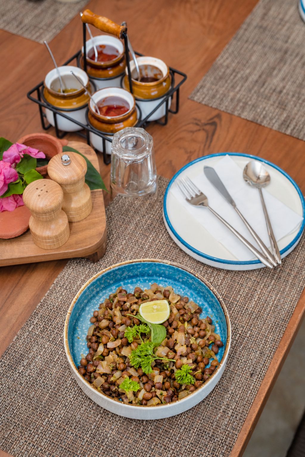 Una mesa de comedor con un tazón azul de garbanzos, un plato con cubiertos y una bandeja con condimentos.