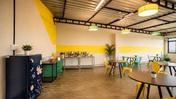 Una cocina moderna con refrigerador, pileta y diversos electrodomésticos. Hay varias mesas y sillas dispuestas para comer.