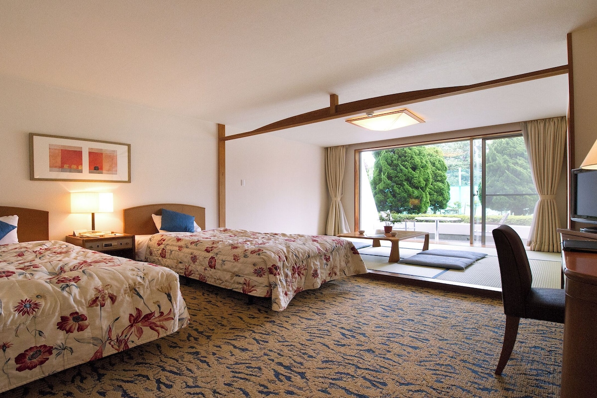 Habitación de hotel con dos camas, un escritorio con televisor y un ventanal con vistas a zonas verdes.