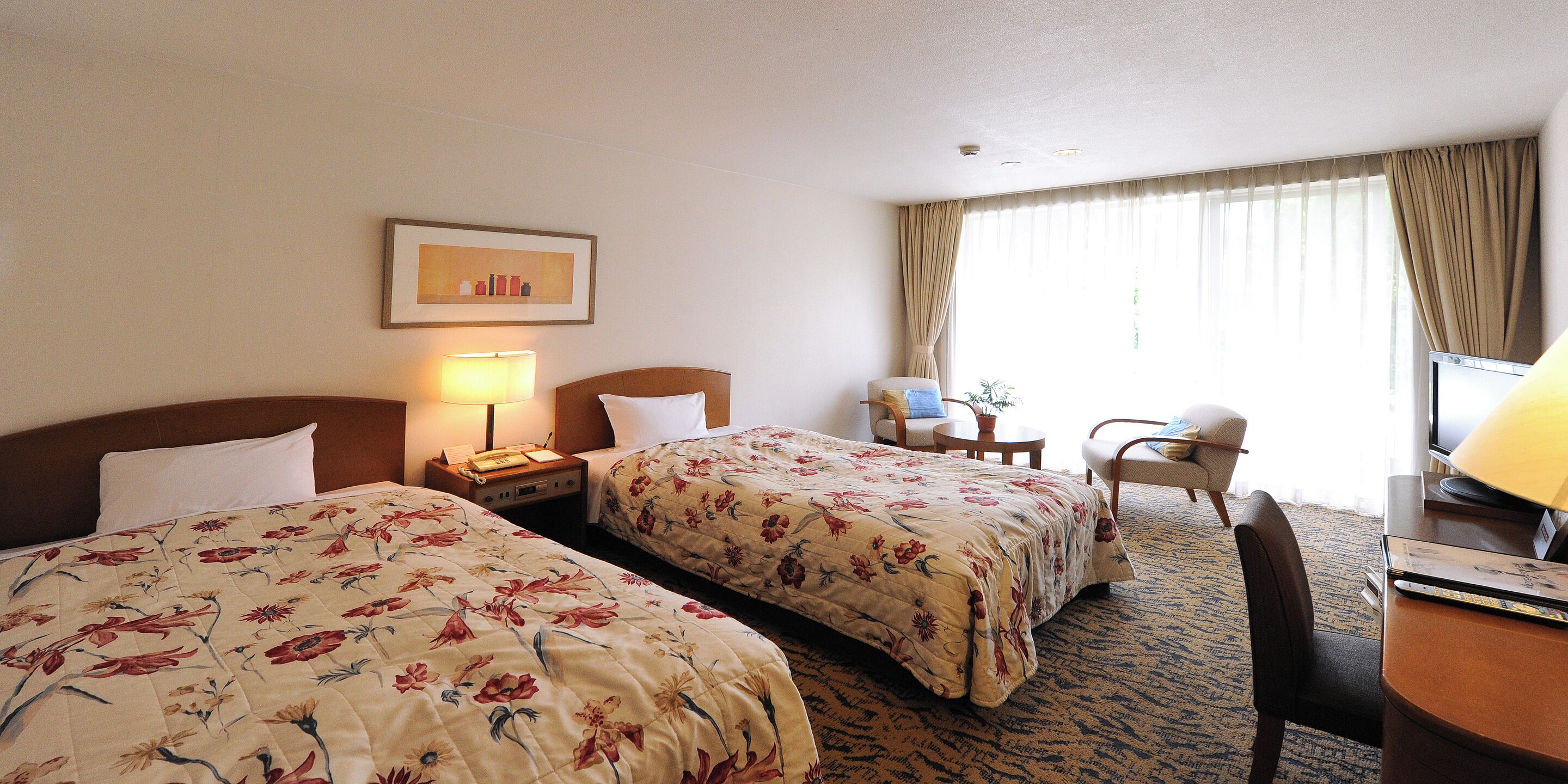 Habitación de hotel con dos camas, un escritorio, una silla, una lámpara y una ventana con cortinas.