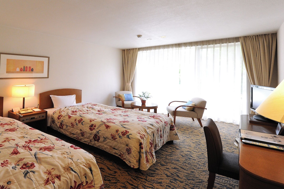 Habitación de hotel con dos camas, un escritorio, una silla, una lámpara y una ventana con cortinas.