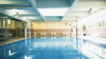 An indoor swimming pool with a clear view of the ceiling structure, a seating area with chairs, and a potted plant.