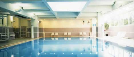 An indoor swimming pool with a clear view of the ceiling structure, a seating area with chairs, and a potted plant.