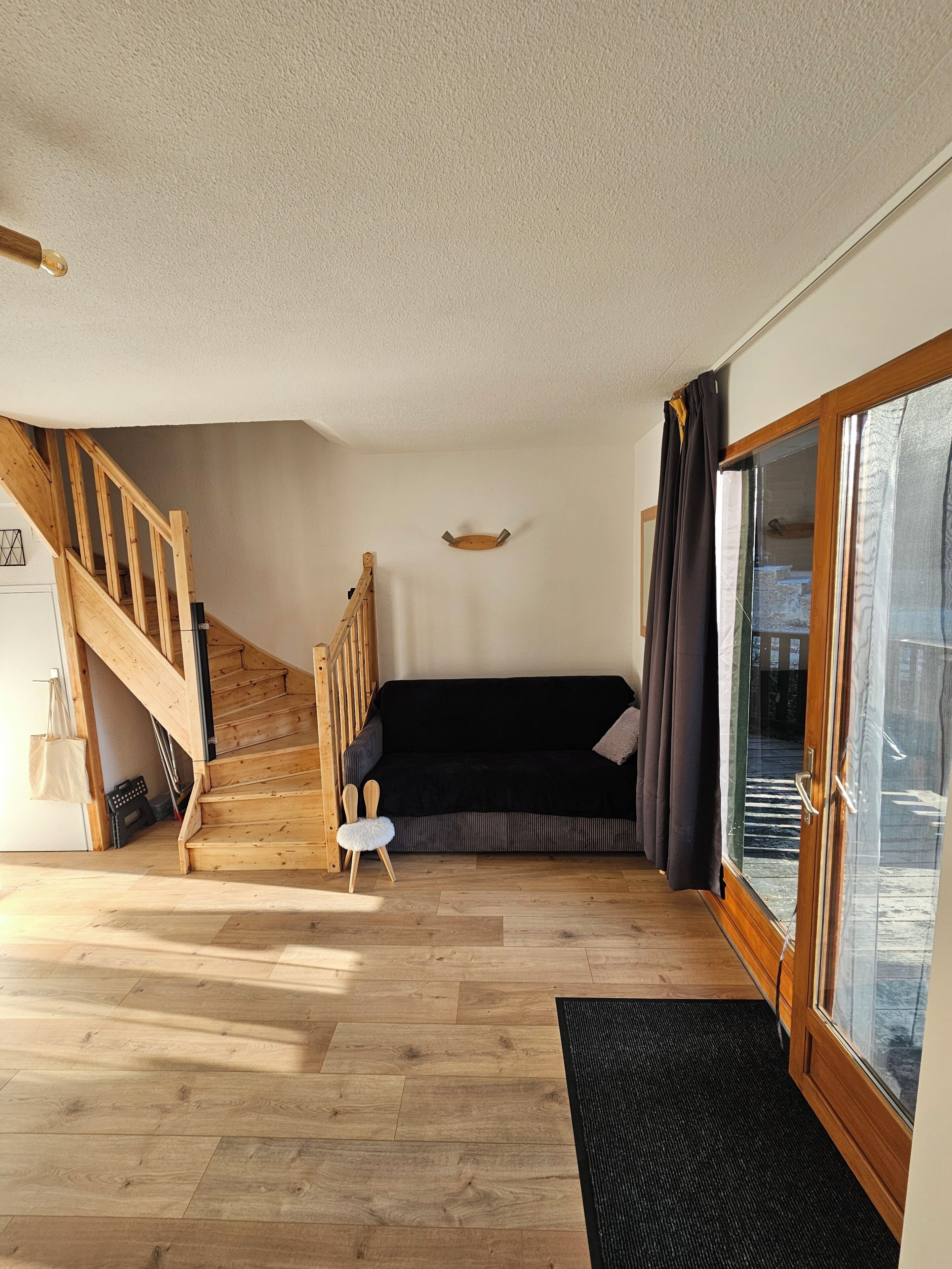 A room with a wooden staircase, a black sofa, a small white chair, and a glass door leading outside.