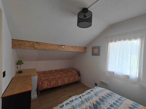 Ein Schlafzimmer mit schrägem Dach, einem Bett mit gemustertem Überwurf, einem kleinen Tisch mit Lampe und einem Fenster mit durchsichtigen Vorhängen.