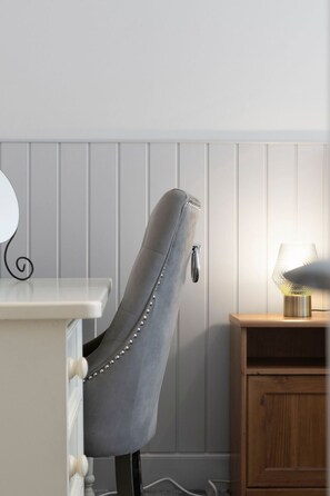 Ein Stuhl mit Metallgestell und Nieten, eine weiße Kommode mit Schubladen sowie ein Nachttisch aus Holz mit einer Lampe.