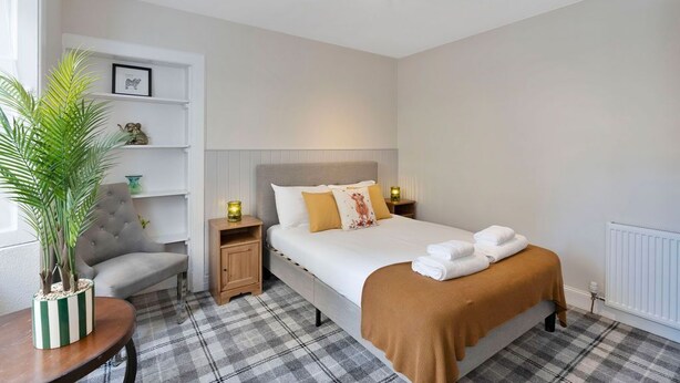 A hotel room with a bed, a chair, a bedside table, a lamp, a shelf with decorative items, and a potted plant. - 2 bedroom property in peaceful Edinburgh location (Edinburgh)