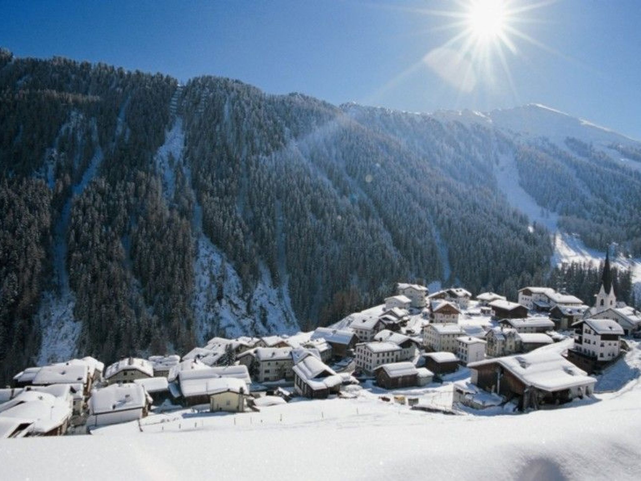 Vila coberta de neve com edifícios e uma igreja, rodeada por montanhas.