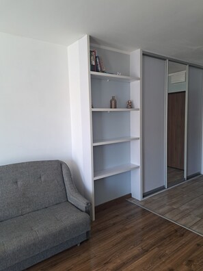 A room with a grey sofa, a white shelving unit with books and decorative items, and a sliding door leading to another room. - OSKAR Studio Apartment (Świnoujście)