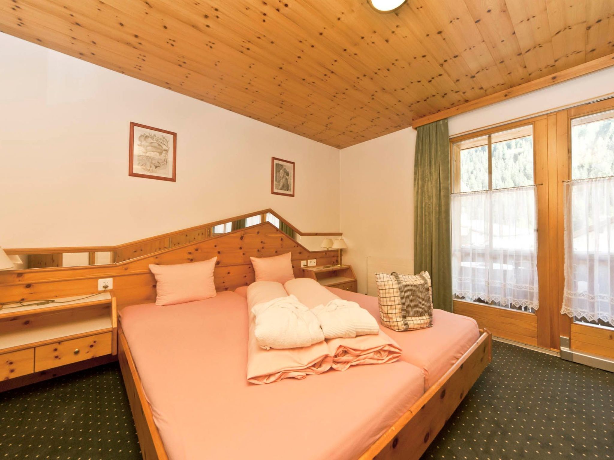 A bedroom with a wooden headboard, a bed with pink bedding, a nightstand, a window with curtains, and two framed pictures on the wall.
