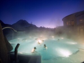 A hot spring area with people relaxing in the water, surrounded by mountains and a building. - Manaröl 607, (Scuol) by Interhome (Scuol)