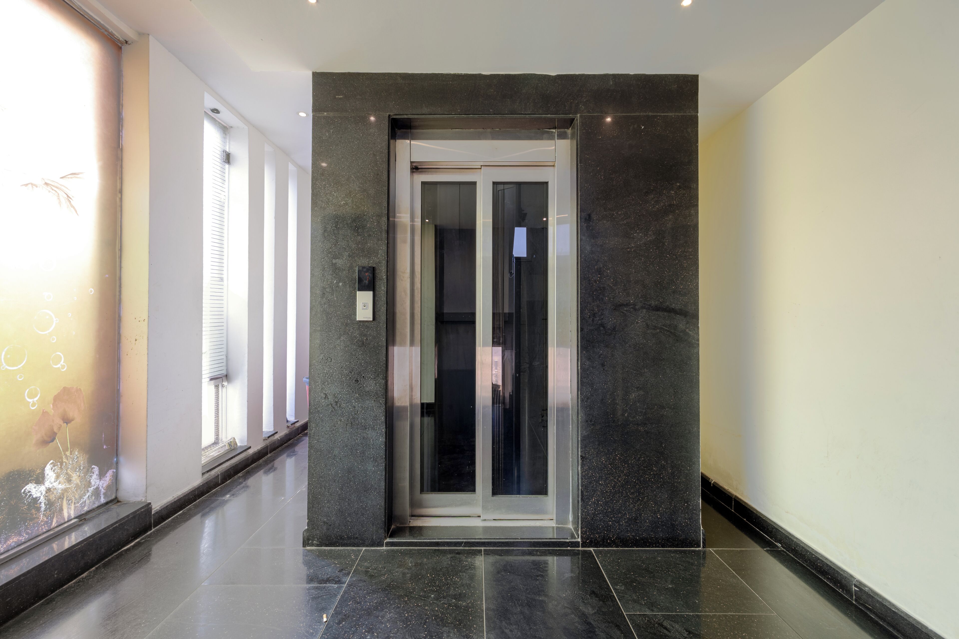Un ascensor moderno con puerta de cristal, ubicado en un pasillo con piso de baldosas y una ventana con persianas.