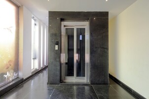 A modern elevator with a glass door, situated in a hallway with a tiled floor and a window with blinds.