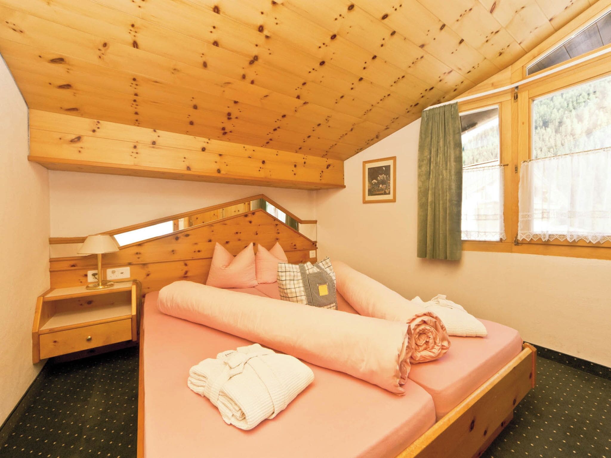 Une chambre confortable avec un plafond en bois, un lit avec une literie rose, une table de chevet, une fenêtre avec des rideaux et un petit tableau accroché au mur.