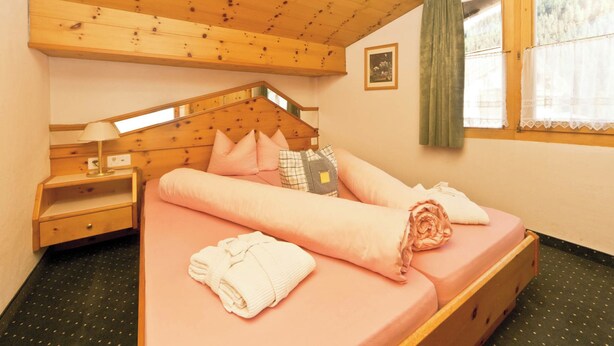 A cozy bedroom with a wooden ceiling, a bed with pink bedding, a nightstand, a window with curtains, and a small picture on the wall. - Appartements Lärchenhof, (Samnaun-Compatsch) by Interhome (Samnaun)