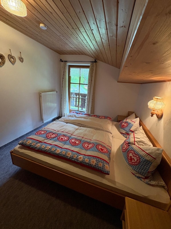 A bedroom with a wooden ceiling, a window with curtains, a radiator, and a bed with patterned bedding.