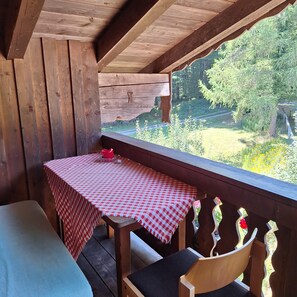 A wooden deck with a table and chairs, a checkered tablecloth, and a view of greenery.