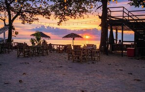 Un restaurant situé en bord de mer, avec une terrasse, un parasol en chaume et une vue sur le coucher de soleil sur l’océan.