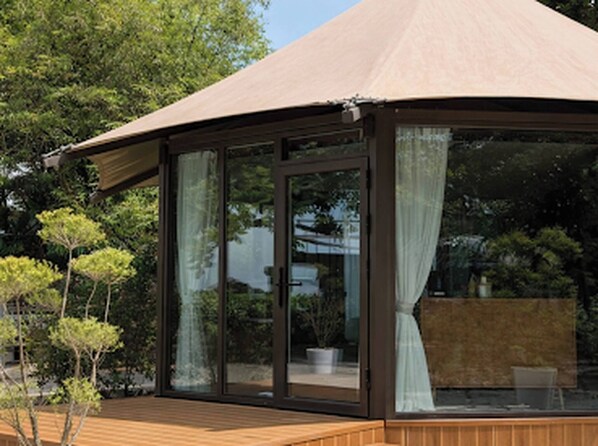 A modern outdoor structure with a glass enclosure, a wooden deck, and a view of surrounding greenery.