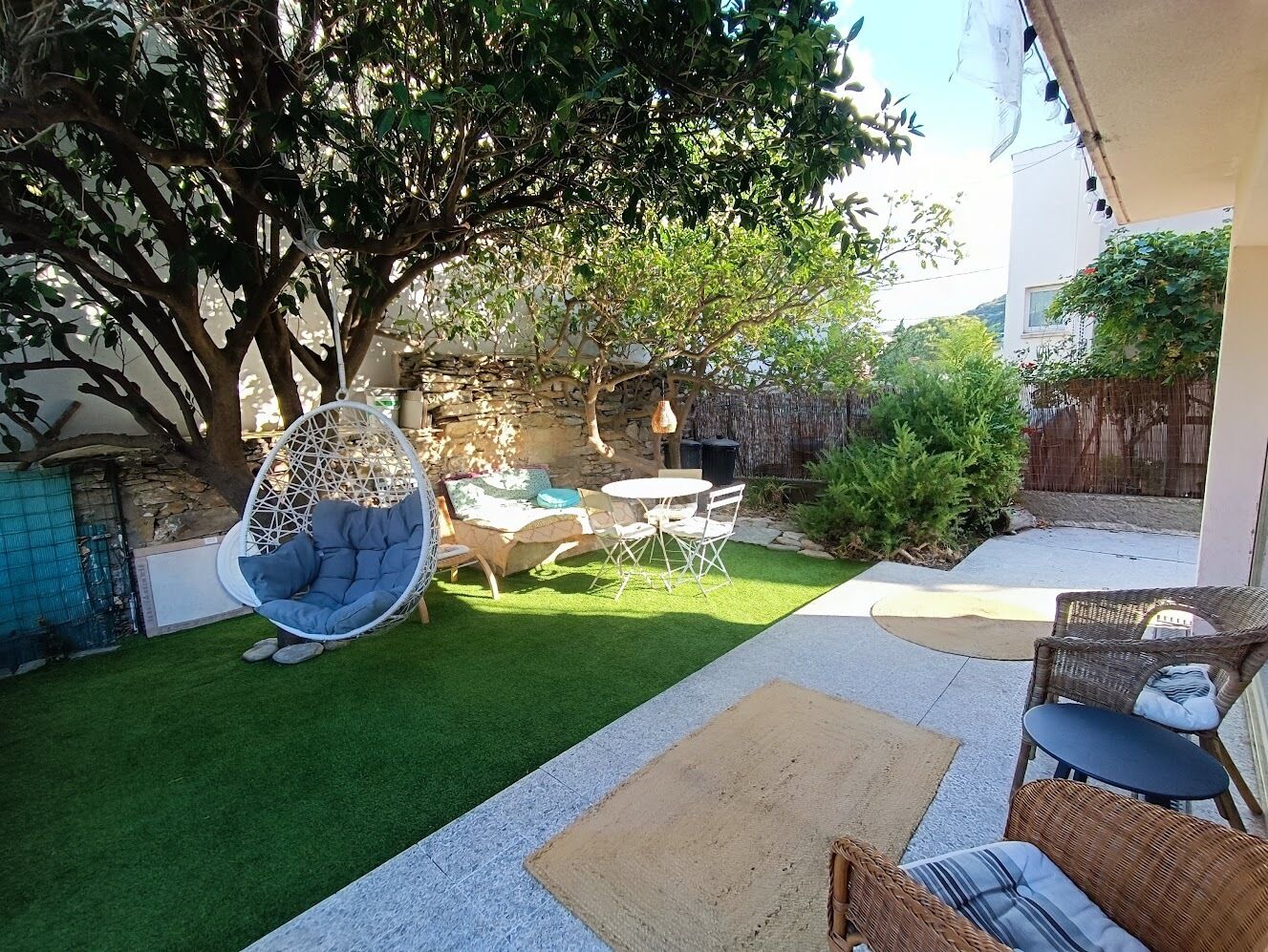 Un jardin avec une pelouse artificielle, un fauteuil suspendu, une table ronde et des meubles en osier.