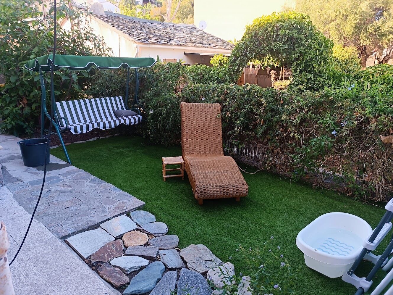 Un patio trasero con un sendero de piedra, una silla lounge de mimbre, un banco a rayas y un taburete de madera pequeño.