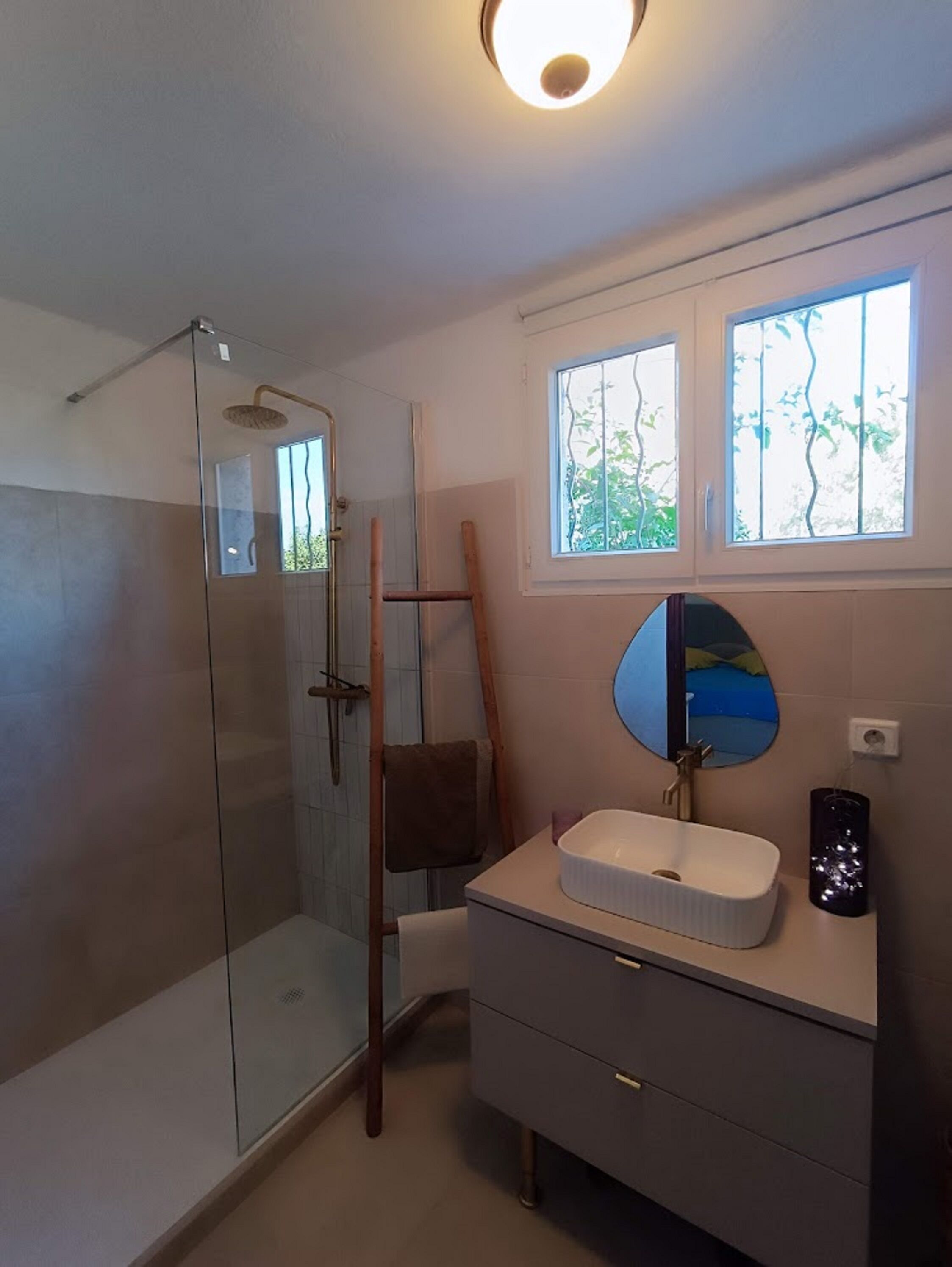 A bathroom with a glass shower enclosure, a wooden ladder, a sink with a mirror, and a window with a view of trees.