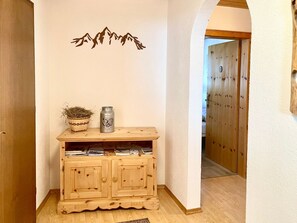 Un mueble de madera con un recipiente metálico y una cesta tejida encima, una pared con silueta de montaña, y una puerta de madera al fondo.