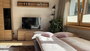 Quarto com cama, televisão, prateleira com objetos decorativos e janela com cortinas.