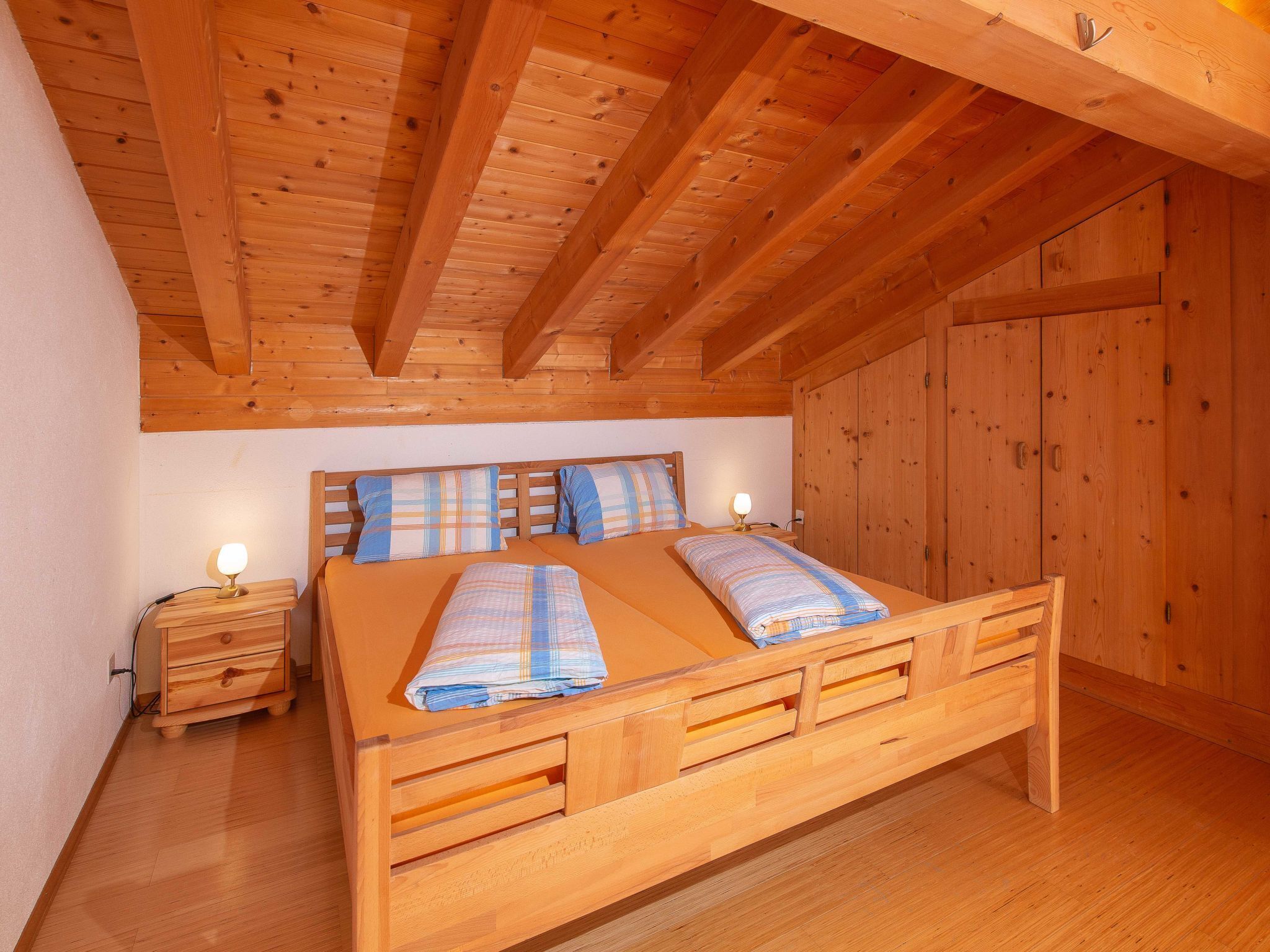 A wooden bed with a headboard, a nightstand, and a wardrobe in a room with wooden beams on the ceiling.