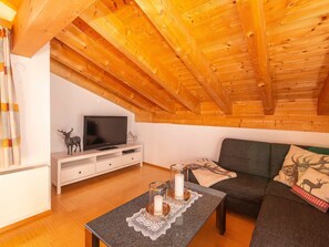 A cozy loft-style room with a wooden ceiling, a flat-screen TV on a white TV stand, a gray sofa, and a small table with candles.