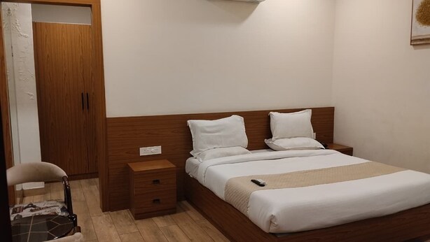 Habitación de hotel con cama de madera, ropa de cama blanca, una mesita de noche, una silla y un equipo de aire acondicionado.