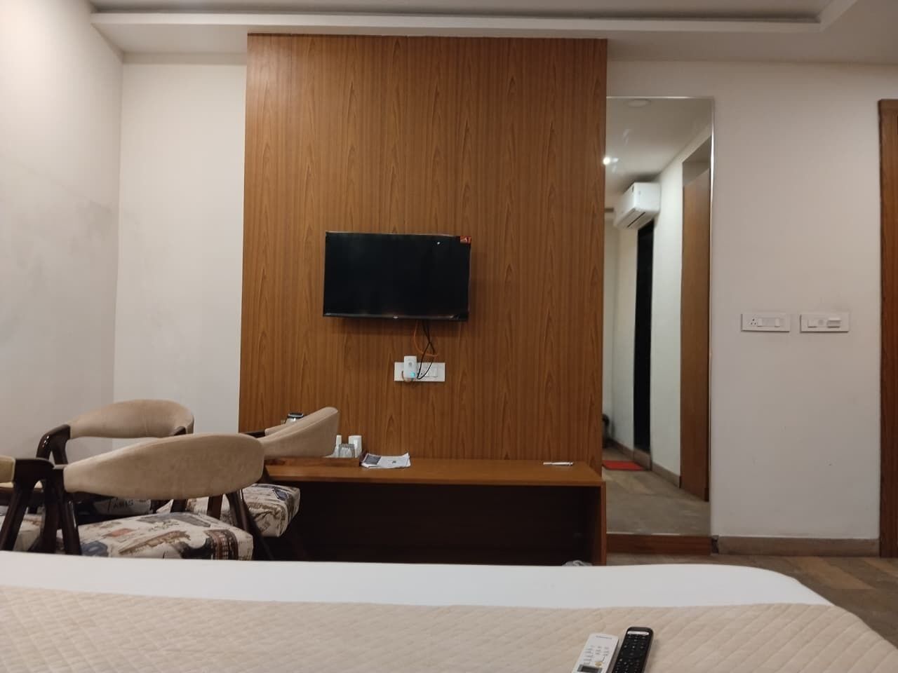 Una habitación de hotel con una cama, un escritorio, una silla, un televisor montado en un panel de madera y una puerta que da a otra habitación.