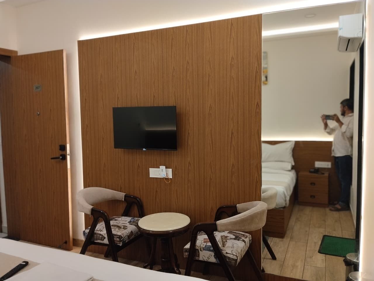 Habitación de hotel con televisión montada en la pared, cama con ropa blanca, mesita de noche y una pequeña mesa redonda con dos sillas.
