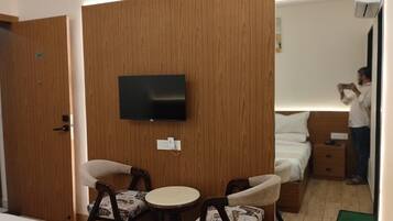 Habitación de hotel con televisión montada en la pared, cama con ropa blanca, mesita de noche y una pequeña mesa redonda con dos sillas.