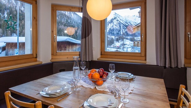 A dining area with a wooden table set for four, surrounded by chairs, with a bowl of fruit and wine glasses on the table.