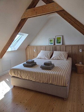 Ein Schlafzimmer mit schrägverdeckter Decke, Holzbalken, einem Bett mit gestreifter Bettwäsche, zwei Kissen, einem Nachttisch und einem kleinen Hocker.