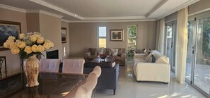 A spacious living room with a glass coffee table, two sofas, and a dining area with a vase of flowers. - Large Luxury Family Home in Upmarket Suburb (Cape Town)