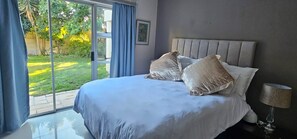 A bedroom with a large bed, a nightstand, a lamp, and a view of a garden through a sliding door. - Large Luxury Family Home in Upmarket Suburb (Cape Town)