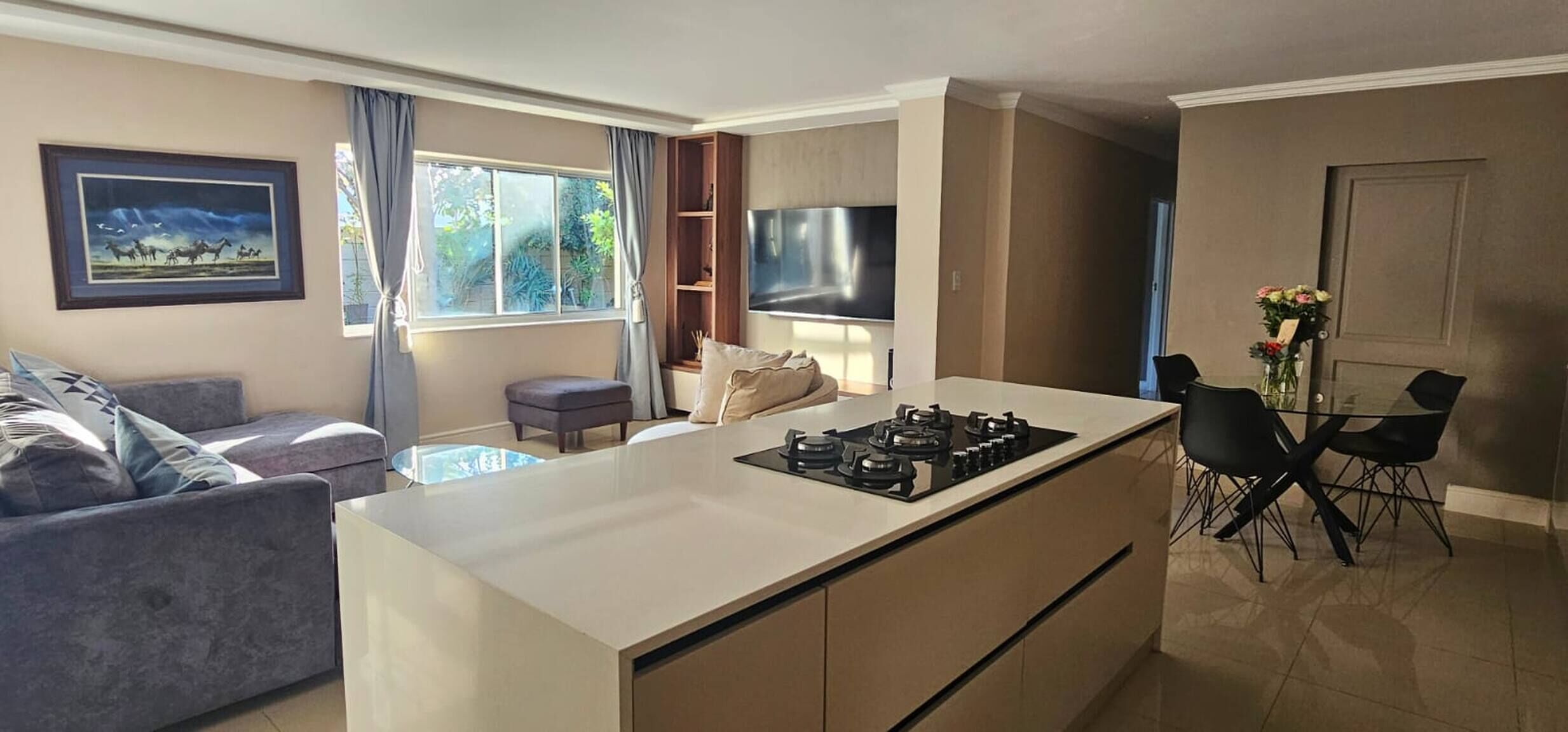A modern kitchen with a central island, dining area with chairs, a sofa, and a TV mounted on the wall.