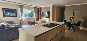 A modern kitchen with a central island, dining area with chairs, a sofa, and a TV mounted on the wall. - Large Luxury Family Home in Upmarket Suburb (Cape Town)