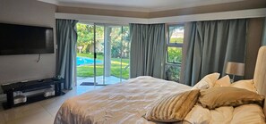 A bedroom with a bed, a TV, a desk, and a view of a swimming pool and greenery. - Large Luxury Family Home in Upmarket Suburb (Cape Town)