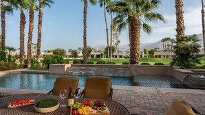 Un espace de restauration aménagé au bord de la piscine, avec une table dressée pour deux personnes, entouré de palmiers et offrant une vue sur un terrain de golf.