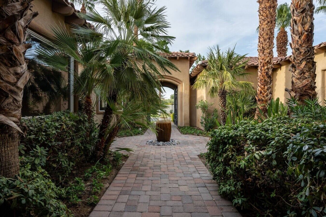 A paved pathway flanked by lush greenery and palm trees, leading to a building with a tiled roof.