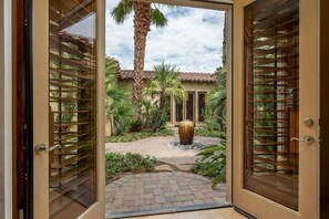 A view through a glass door into a courtyard with palm trees, a large vase, and a tiled pathway. - Canyon House at PGA West | Poolside Golf Views (La Quinta)