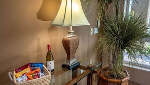 Une pièce avec une table en verre, une lampe, une bouteille de vin, un panier de collations et une plante en pot.