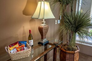 Ein Zimmer mit einem Glastisch, einer Lampe, einer Weinflasche, einer Snackkörbchen und einer Blumentopf.