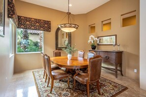 A dining area with a round wooden table, leather chairs, a chandelier, a decorative mirror, and a view of greenery outside.