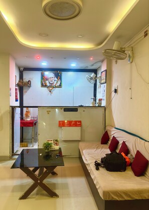 Habitación de hotel con una recepción, una cama con almohadas rojas, una mesa con una planta y un ventilador de techo.